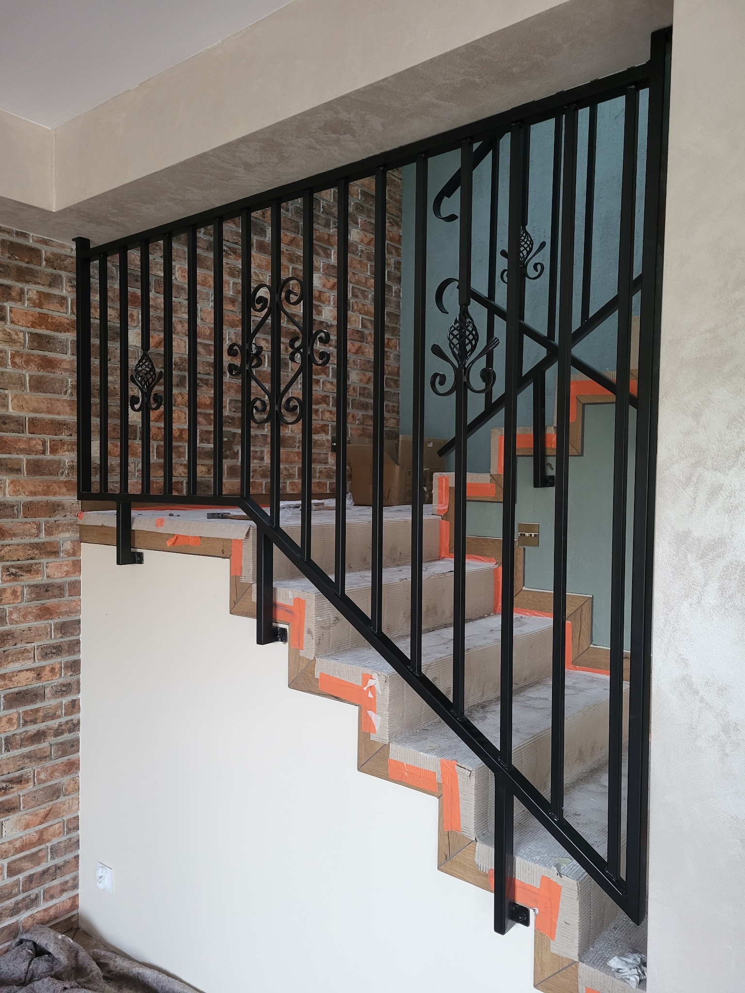 Czarna, metalowa balustrada schodowa z dekoracyjnymi elementami, zamontowana na zabezpieczonych schodach wewnątrz budynku. Widoczna ceglana ściana i jasne ściany.