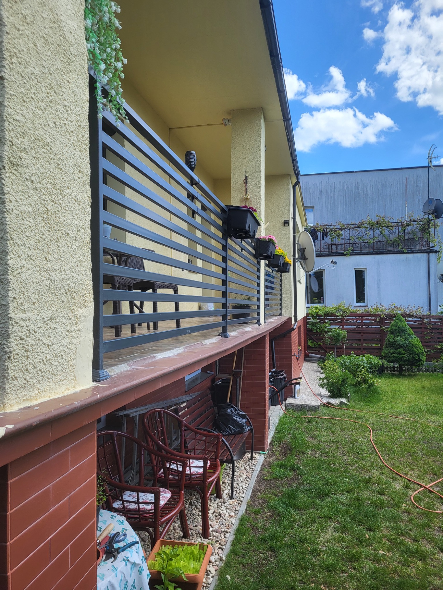 Nowoczesna, pozioma balustrada balkonowa w kolorze antracytu, z widokiem na meble ogrodowe i zadbany trawnik. Elementy dekoracyjne: kwiaty w donicach.