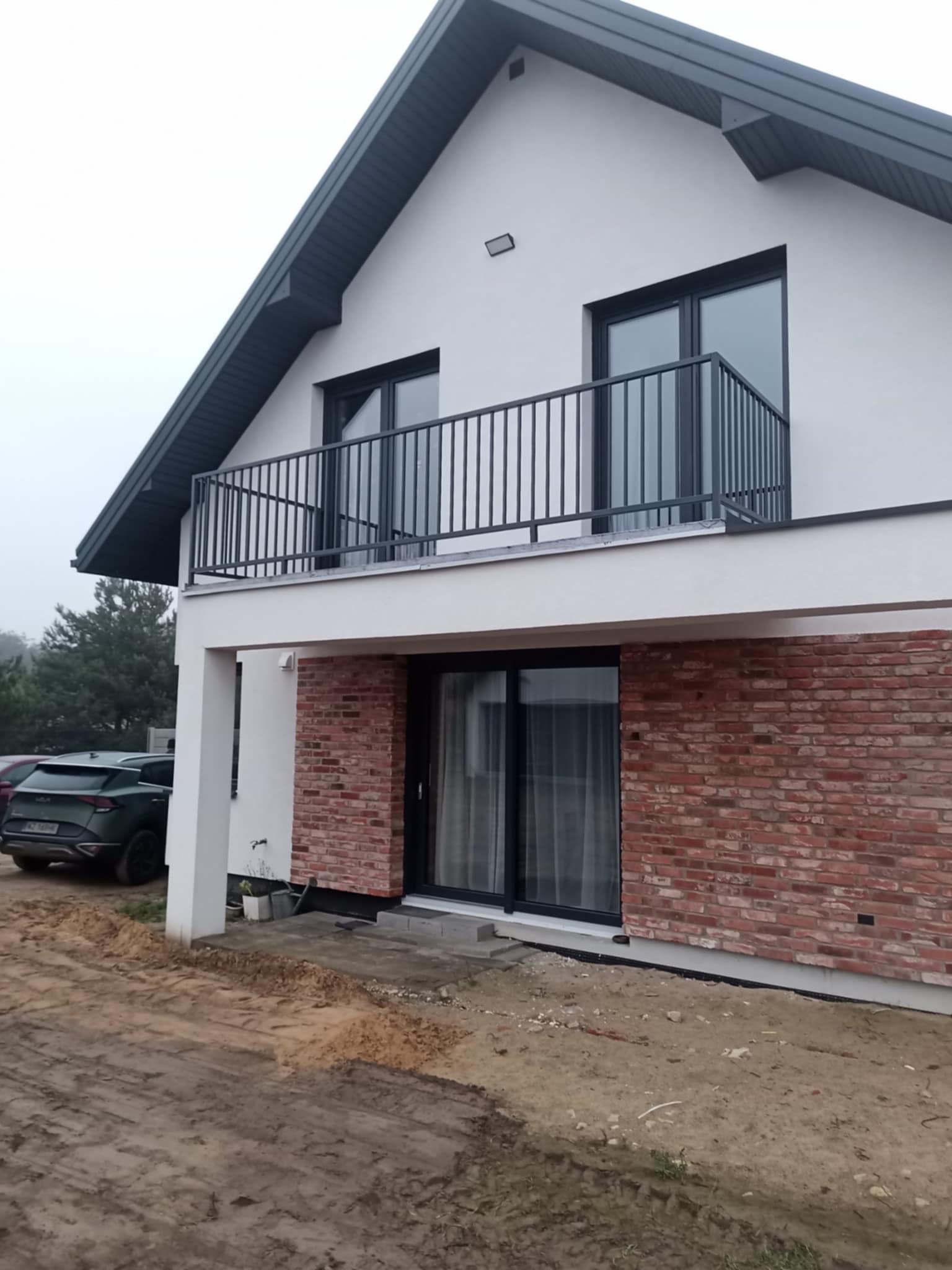 Nowoczesny dom z balkonem i czarną balustradą. Elewacja z białego tynku i cegły klinkierowej. Widoczny fragment podjazdu i samochód. Ujęcie z dołu, pochmurny dzień.