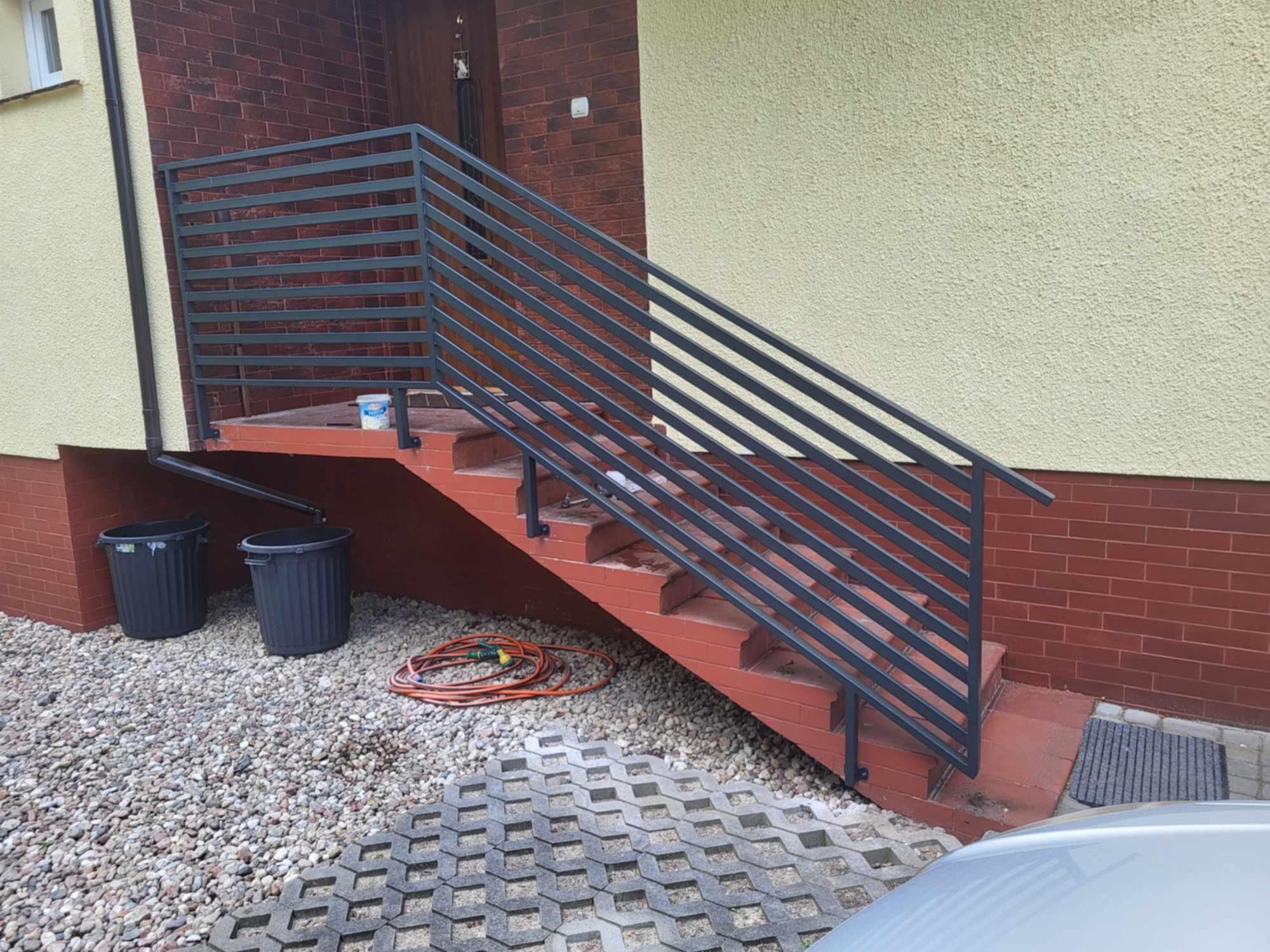 Nowoczesna, pozioma balustrada schodowa z metalu w kolorze grafitowym, zamontowana przy wejściu do domu z ceglaną elewacją i beżowym tynkiem.