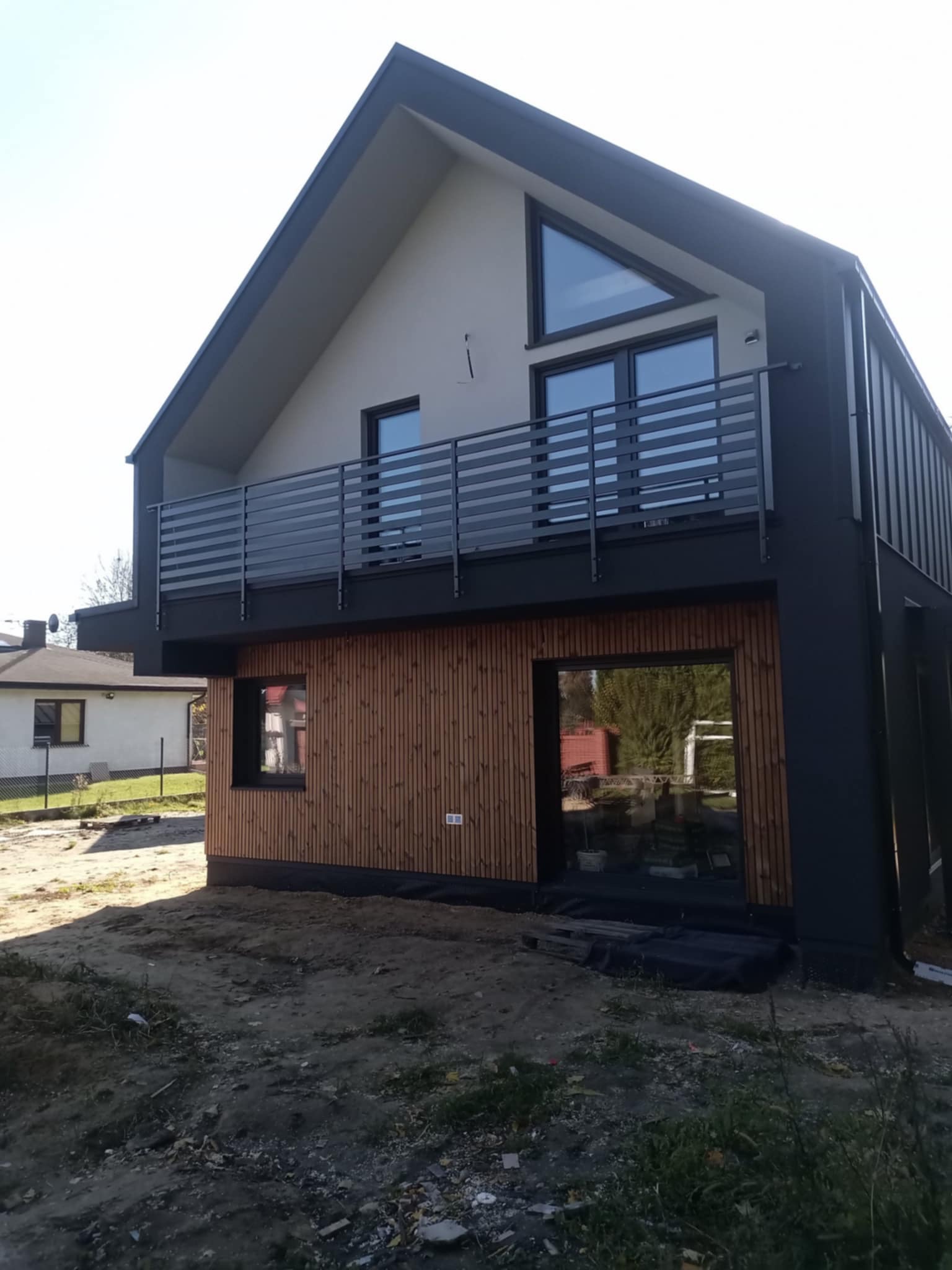 Nowoczesny dom z drewnianą elewacją i szarą balustradą balkonową. Widoczne okna o nietypowym, trójkątnym kształcie. Surowe otoczenie.