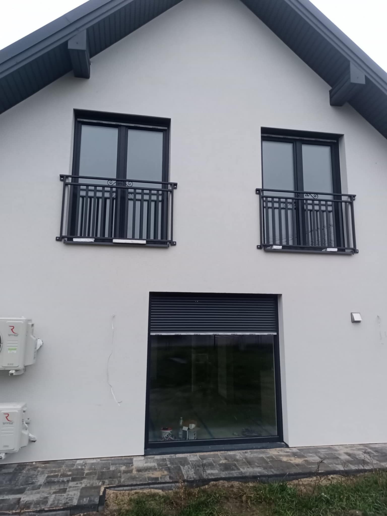 Elewacja domu z dwoma oknami z czarnymi balustradami balkonowymi o dekoracyjnym wzorze i oknem z roletą. Widoczne klimatyzatory i fragment chodnika.