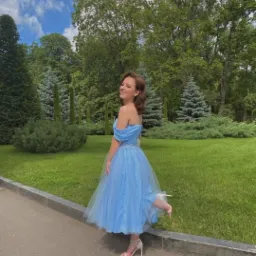 Uśmiechnięta młoda kobieta w błękitnej tiulowej sukience bez ramiączek i białych szpilkach, pozuje na tle parku w słoneczny dzień.