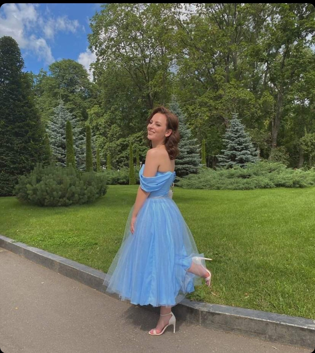 Uśmiechnięta młoda kobieta w błękitnej tiulowej sukience bez ramiączek i białych szpilkach, pozuje na tle parku w słoneczny dzień.