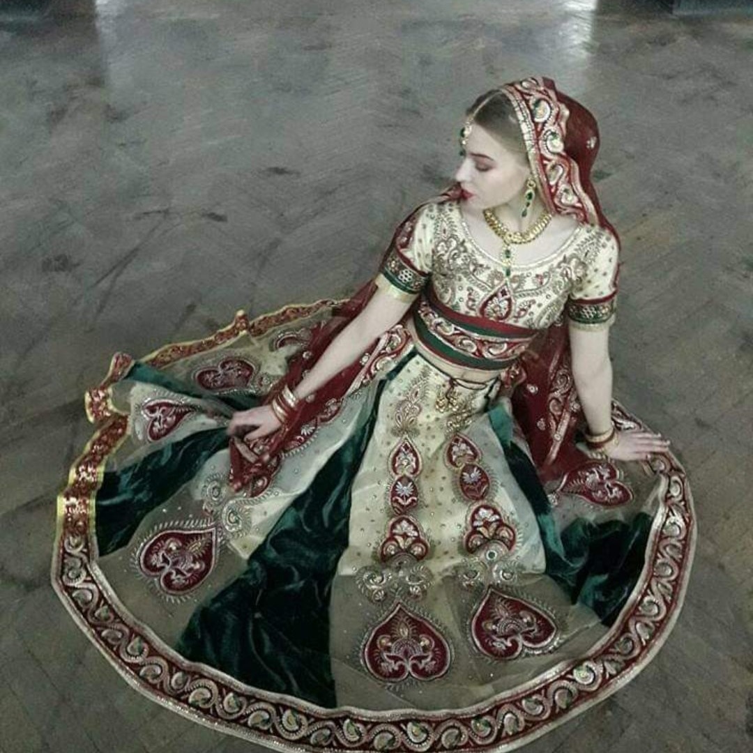 Kobieta siedzi na podłodze w bogato zdobionej, tradycyjnej sukni indyjskiej z zielonymi i kremowymi panelami, z misternymi haftami i biżuterią, patrząc w lewo.