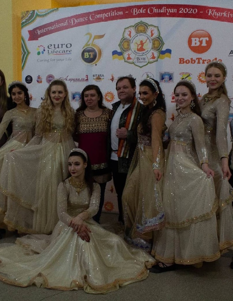 Grupa tancerzy w błyszczących, beżowych strojach z ozdobnymi lamówkami pozuje na tle plakatu 'International Dance Competition - Bole Chudiyan 2020 - Kharkiv'.