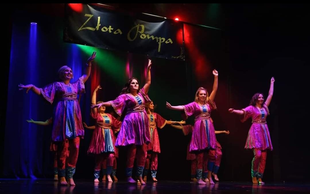 Zespół taneczny w fioletowych kostiumach z niebieskimi wstawkami występuje na scenie pod banerem z napisem 'Złota Pompa', oświetlony kolorowymi reflektorami.