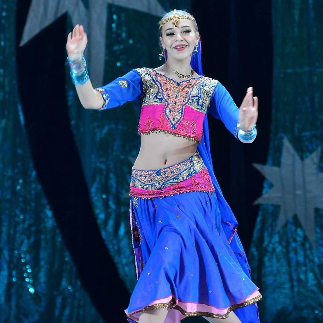 Młoda kobieta w niebiesko-różowym, bogato zdobionym stroju tanecznym, z bransoletkami i nakryciem głowy, uśmiecha się i tańczy na tle niebieskiej sceny.