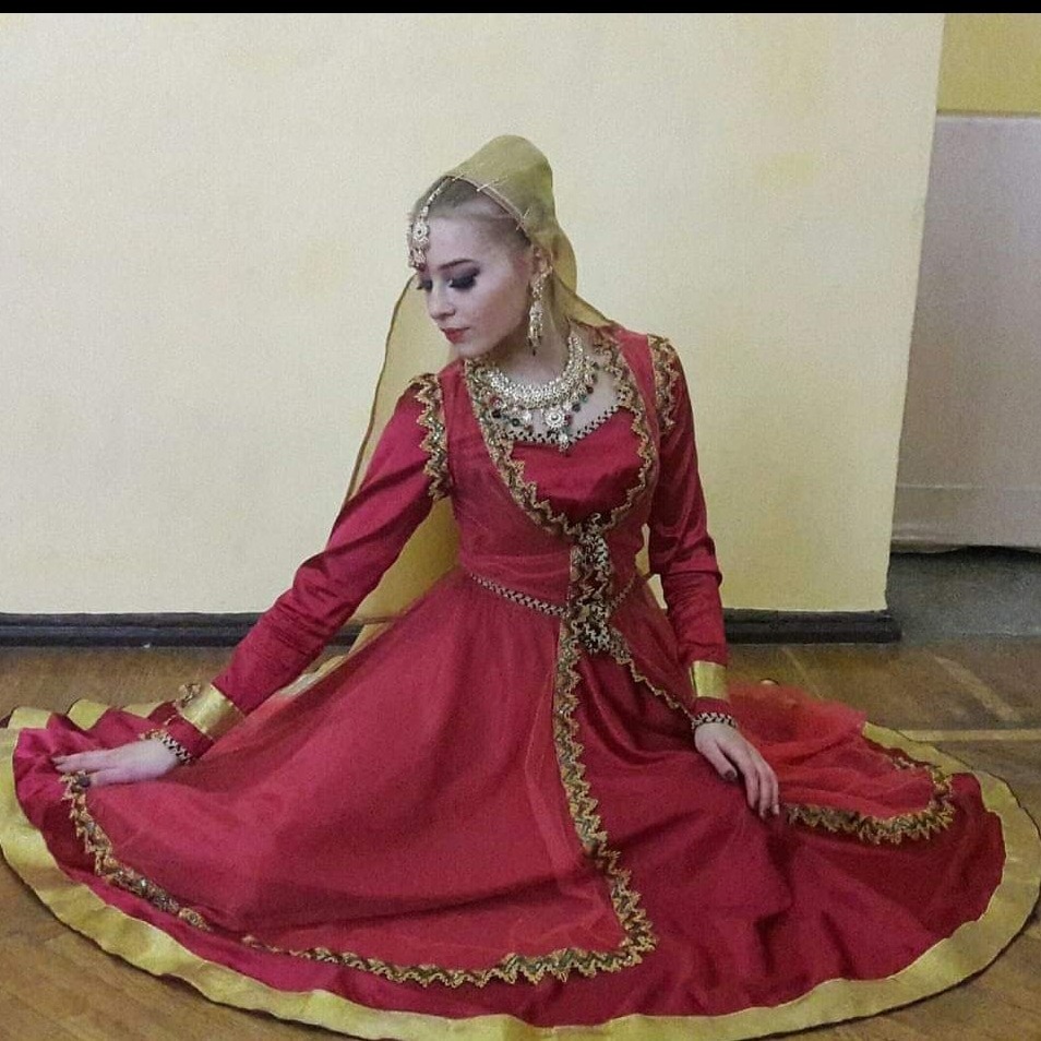 Młoda kobieta w czerwonej, bogato zdobionej sukni z elementami złota i nakryciem głowy, siedząca na podłodze. Suknia o kroju balowym, z długimi rękawami i szeroką spódnicą, uzupełniona jest...