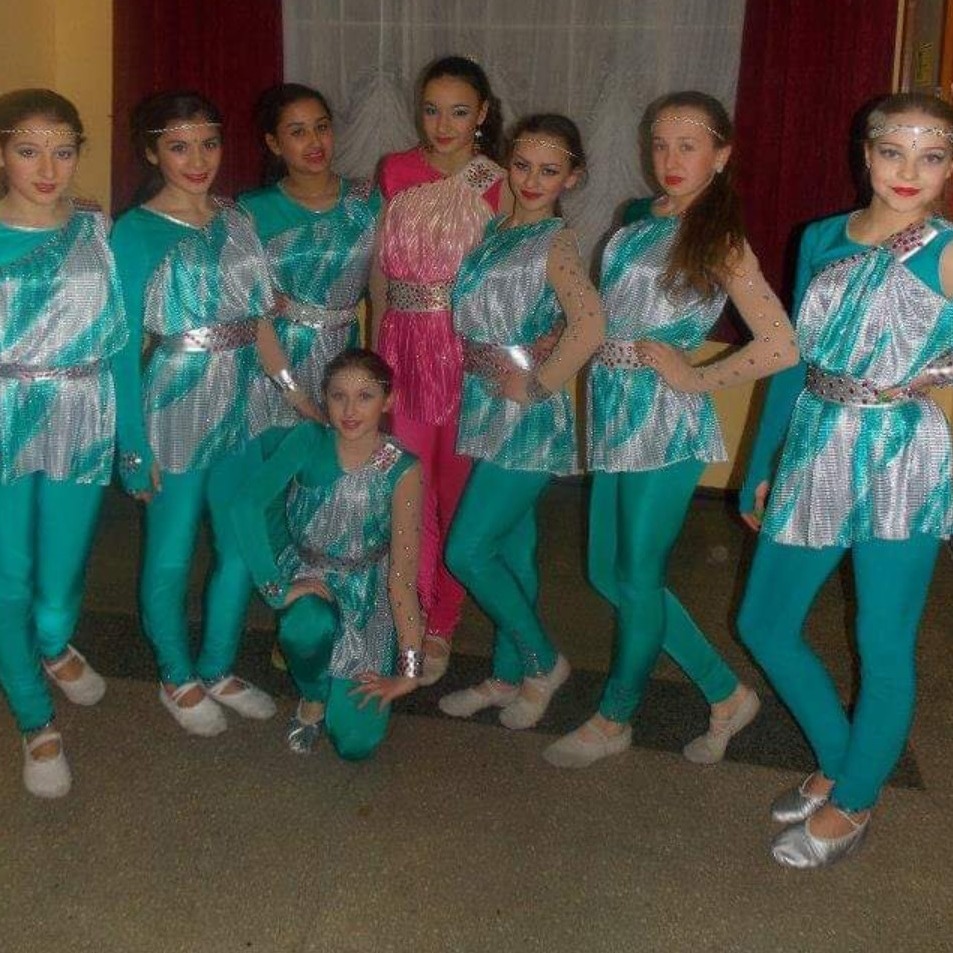 Zespół tancerek w turkusowo-srebrnych kostiumach z paskami, jedna tancerka w różowym stroju, pozują do zdjęcia przed ciemną zasłoną.