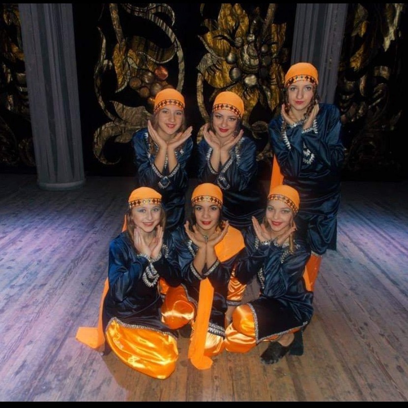 Grupa młodych kobiet w granatowo-pomarańczowych strojach ludowych i czepkach, pozująca na scenie z dekoracyjnym tłem.