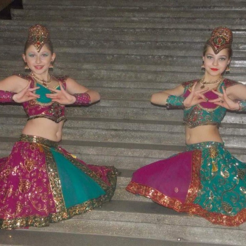 Dwie młode tancerki w bogato zdobionych, kolorowych strojach indyjskich, prezentują układ taneczny na tle betonowych schodów.