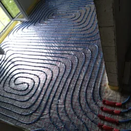 Montaż ogrzewania podłogowego: niebieskie rurki ułożone spiralnie na folii z czerwoną siatką, widoczne zabezpieczenia rur przy ścianie, surowy stan wykończenia.