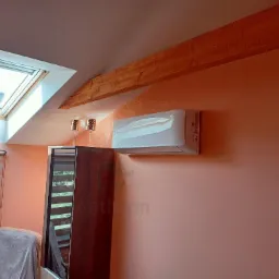 Biały klimatyzator ścienny zamontowany w pokoju na poddaszu z oknem dachowym, widoczna drewniana belka stropowa i rolety okienne.