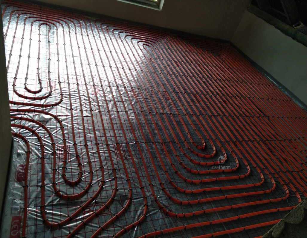 Montaż ogrzewania podłogowego: czerwone rurki ułożone w pętle na folii z siatką, widoczne fragmenty ścian i okna w tle.