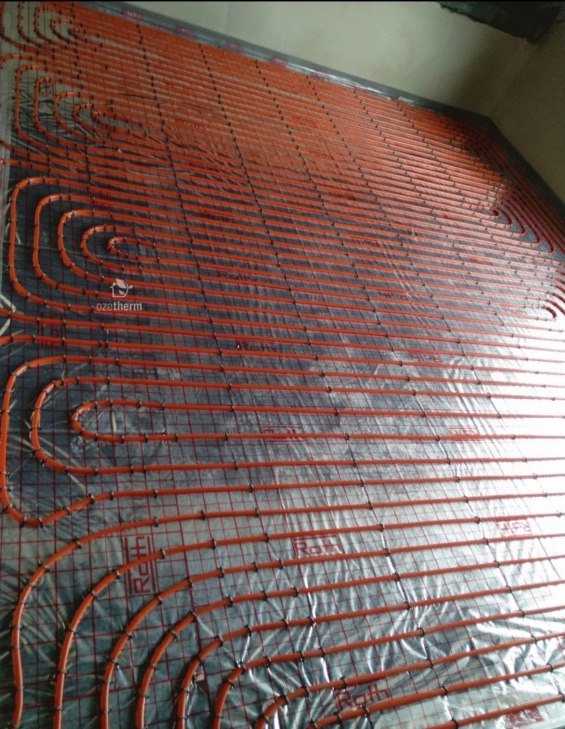 Instalacja ogrzewania podłogowego z rurkami w kolorze pomarańczowym, zamocowanymi na siatce na folii izolacyjnej, widoczne logo producenta rur Roth i częściowo logo Ozetherm.