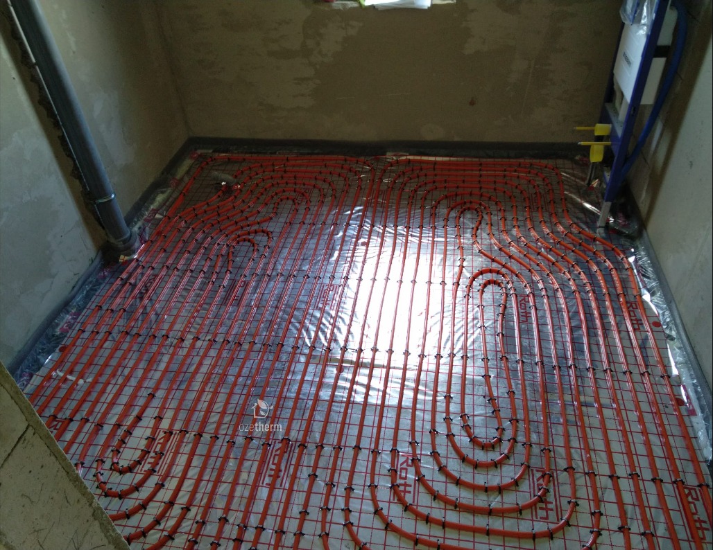 Instalacja ogrzewania podłogowego z czerwonych rur na siatce, widoczna folia izolacyjna i szare ściany w surowym stanie.