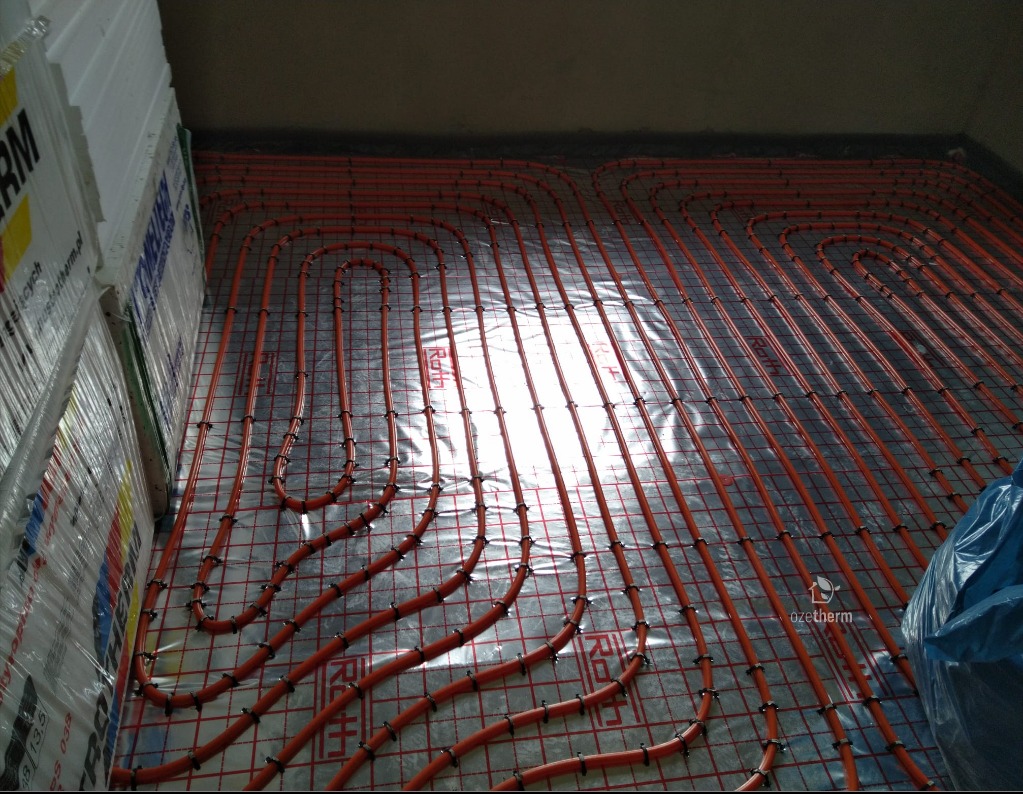 Montaż ogrzewania podłogowego z rur w kolorze pomarańczowym na metalowej siatce, widoczne płyty izolacyjne przy ścianie.