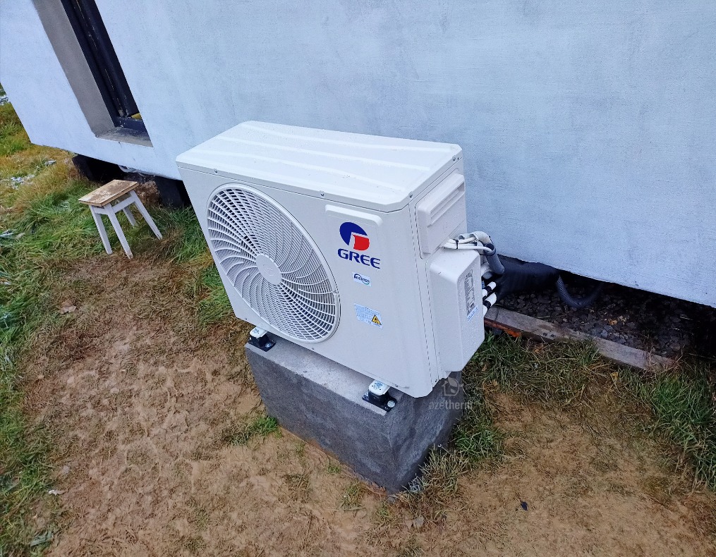 Zewnętrzna jednostka klimatyzacji Gree zamontowana na betonowym fundamencie przy ścianie budynku, widoczny fragment trawnika i mały stołek.
