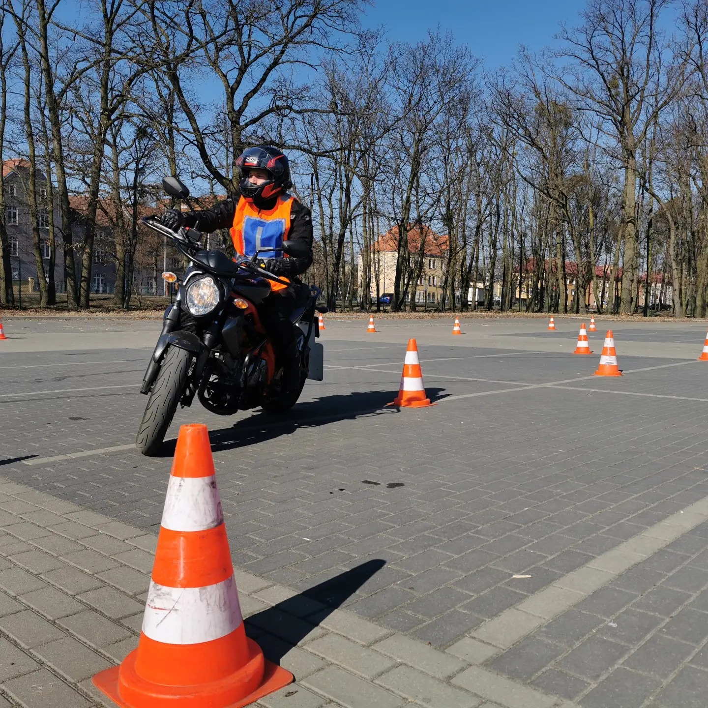 Kursant w kasku i kamizelce z literą 'L' na motocyklu, pokonuje tor przeszkód z pachołkami na placu manewrowym, w tle budynki i drzewa bez liści.