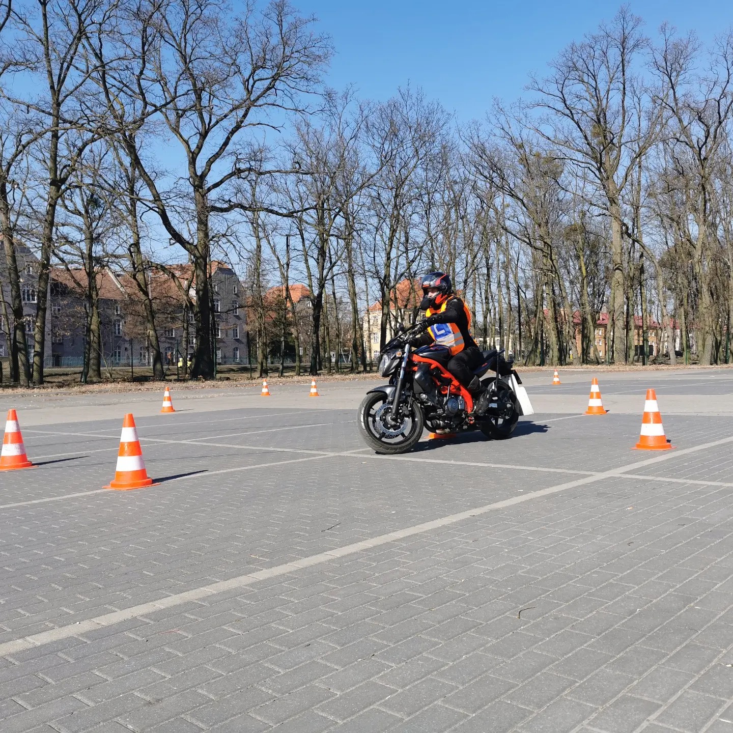 Motocyklista w kamizelce odblaskowej ćwiczy manewry na placu z pachołkami, w tle budynki i drzewa bez liści.