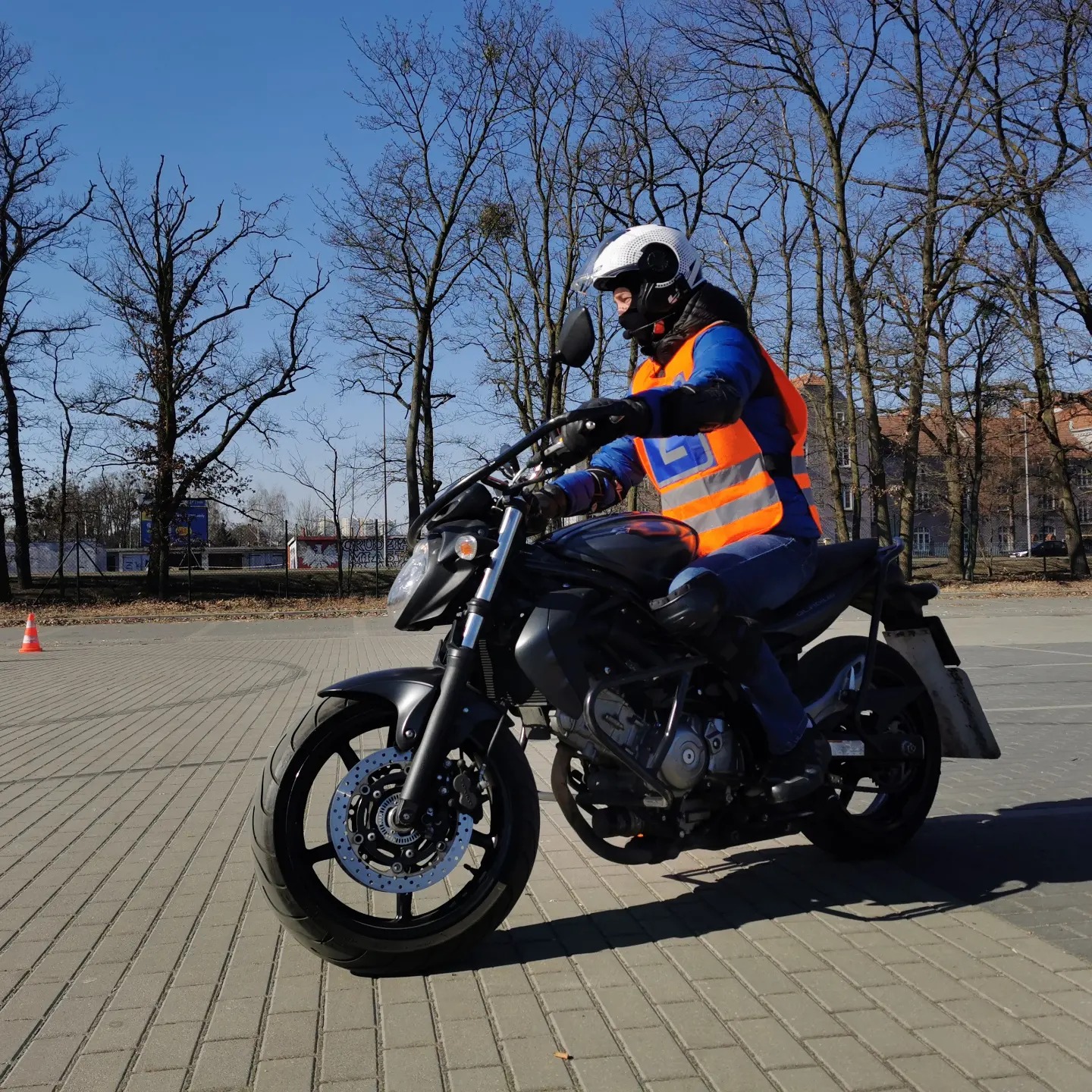 Osoba w kasku i kamizelce odblaskowej na czarnym motocyklu na placu manewrowym z kostki brukowej, drzewa bez liści w tle.