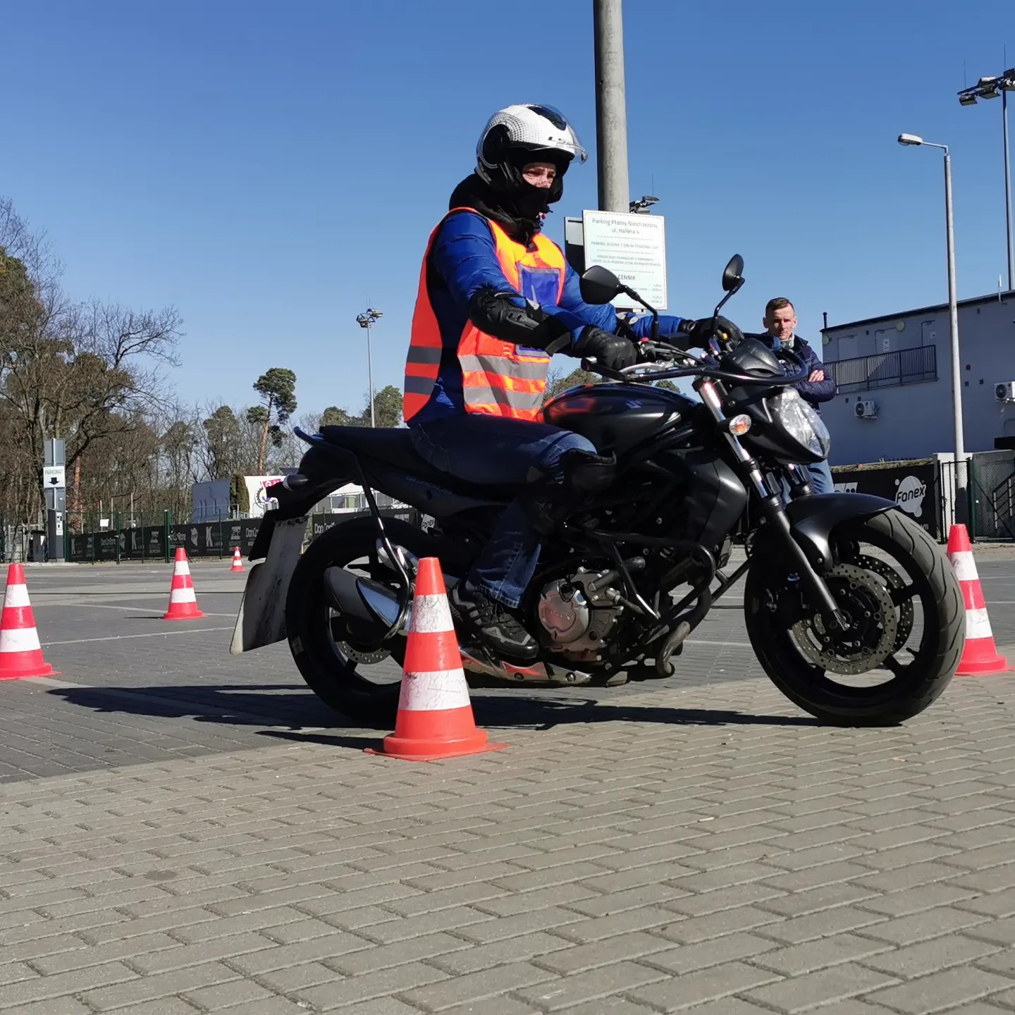 Kobieta w kasku i kamizelce odblaskowej na czarnym motocyklu, ćwicząca manewry na placu z pachołkami; instruktor obserwuje w tle.