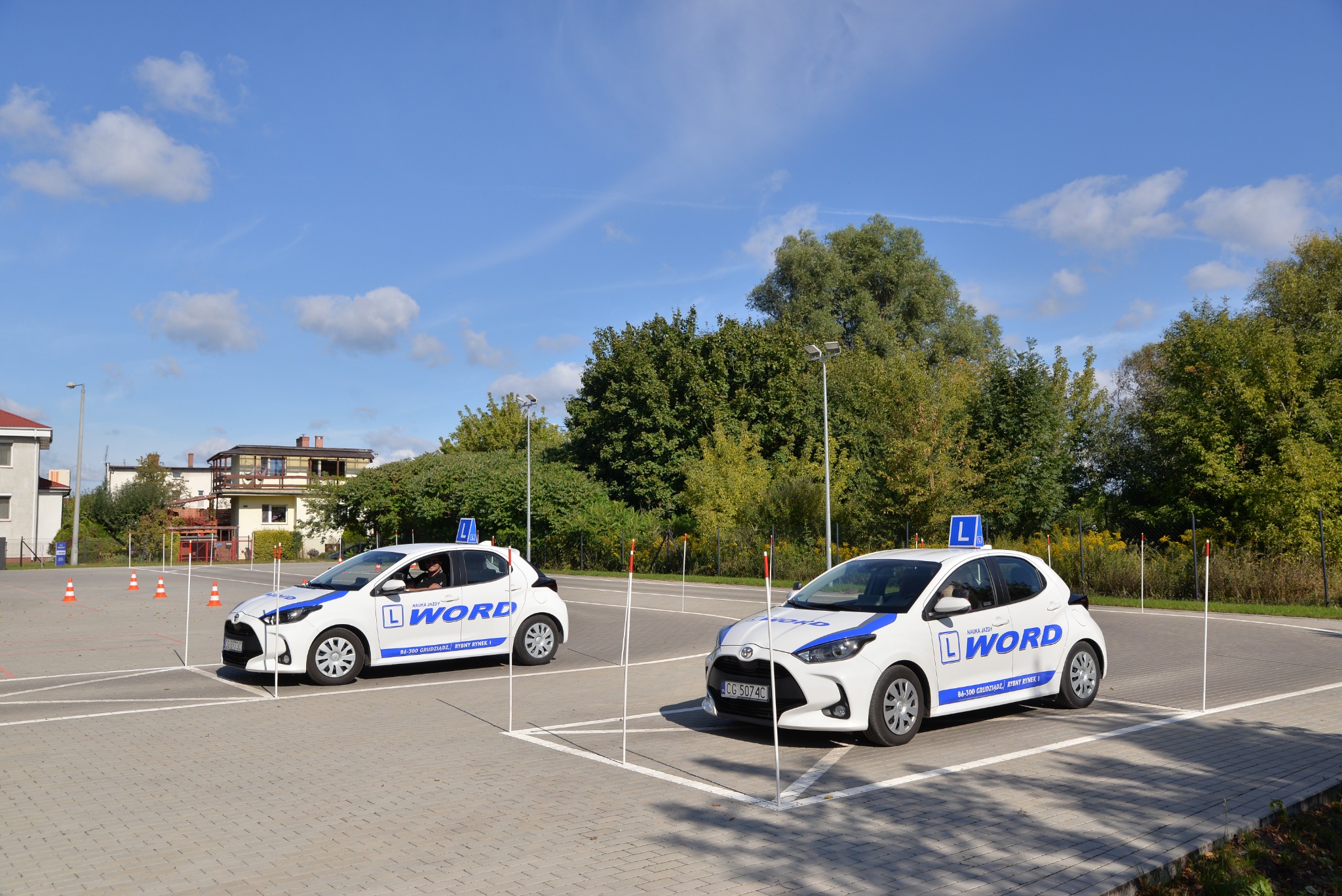 Dwa białe samochody szkoły jazdy WORD na placu manewrowym, ustawione między słupkami, w tle budynek i drzewa pod błękitnym niebem.