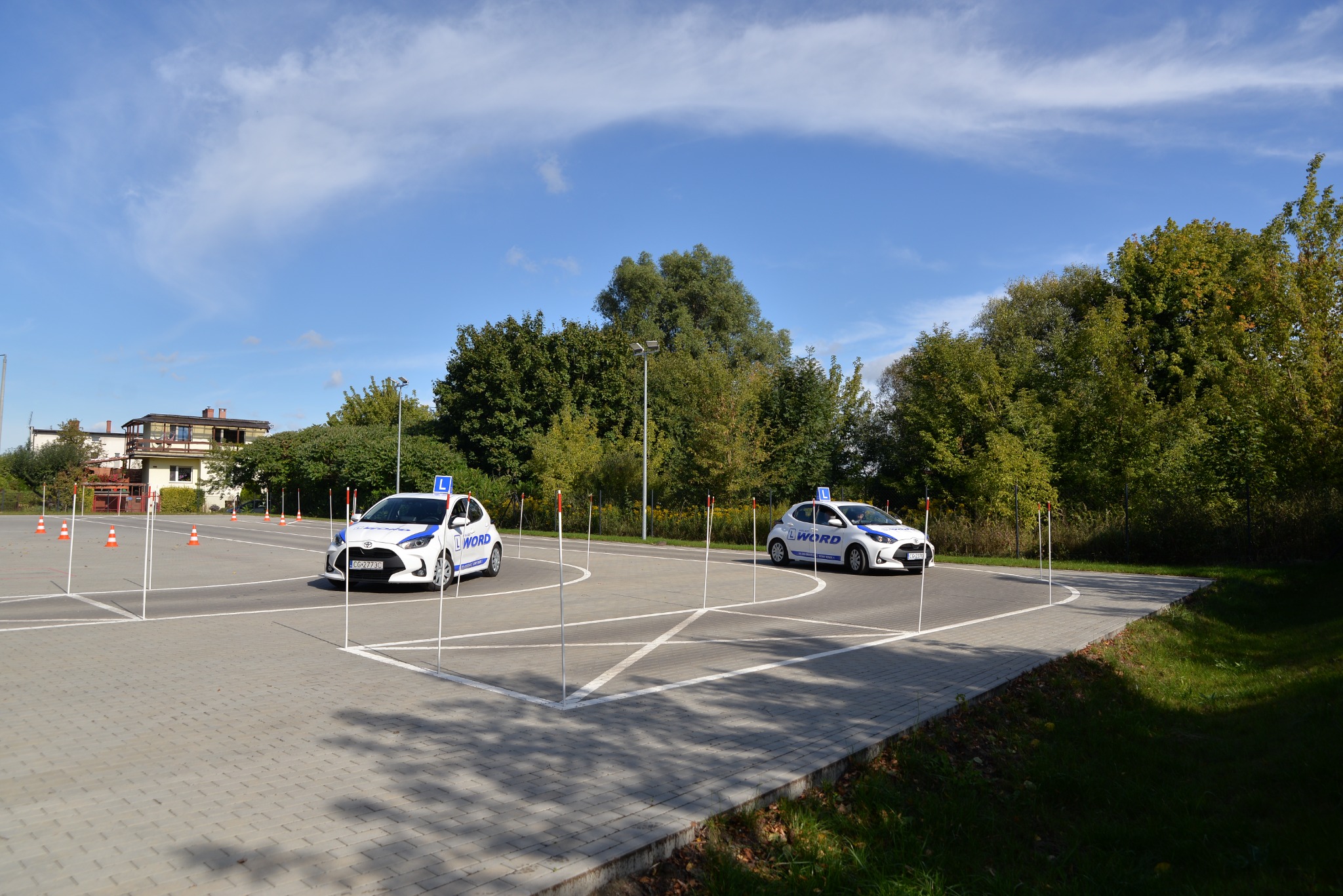 Dwa białe samochody z literą 'L' na dachu, na placu manewrowym z pachołkami i wyznaczonymi liniami, w tle budynek i drzewa, słoneczny dzień.