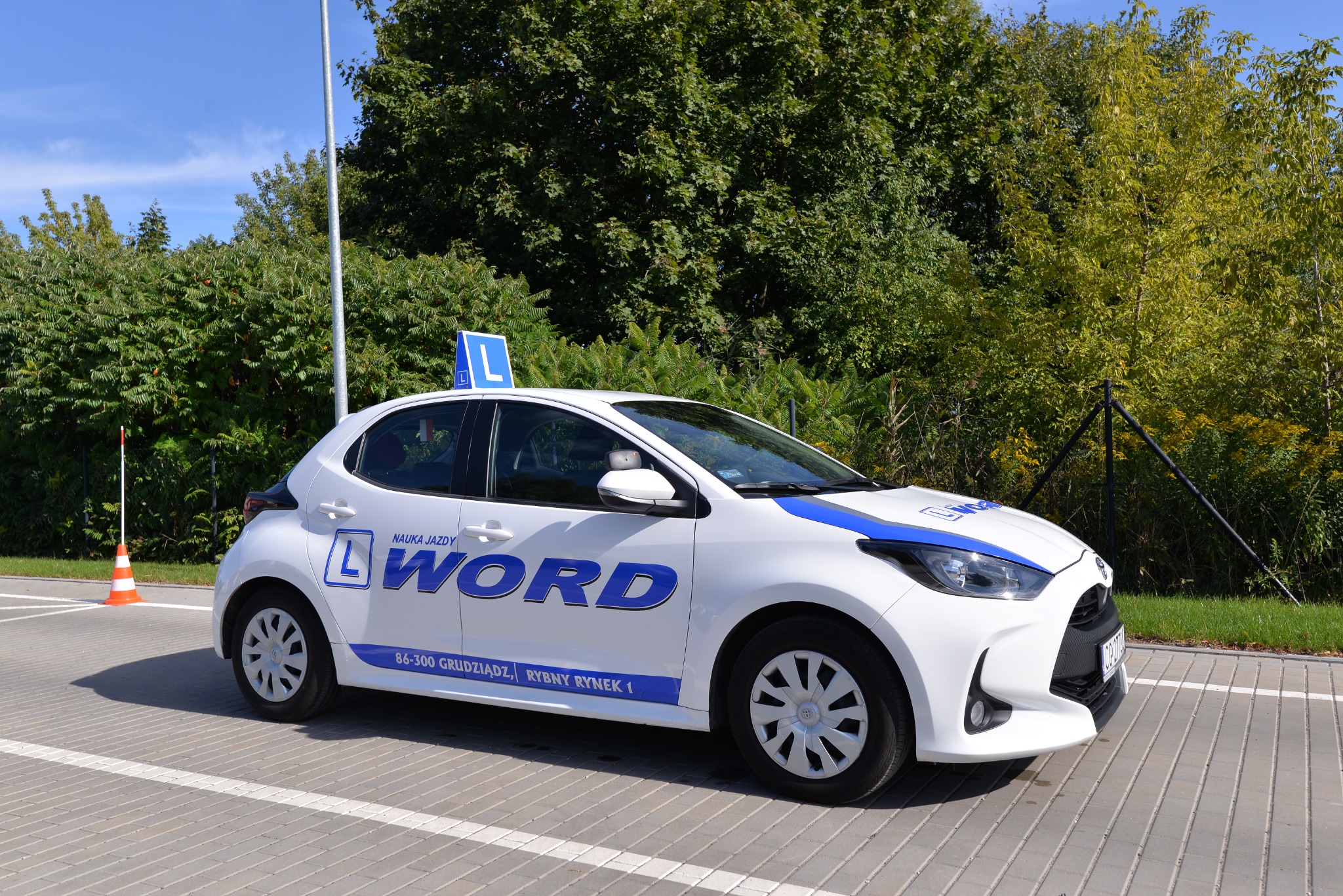 Biały samochód oznaczony logo i napisem firmy szkoleniowej z niebieskim paskiem na dachu i literą 'L' na dachu, zaparkowany na parkingu z kostki brukowej, w tle zieleń drzew i krzewów.