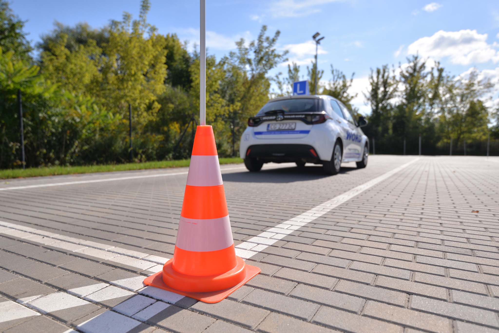 Pomarańczowo-biały pachołek na placu manewrowym z białym autem z literą L w tle.