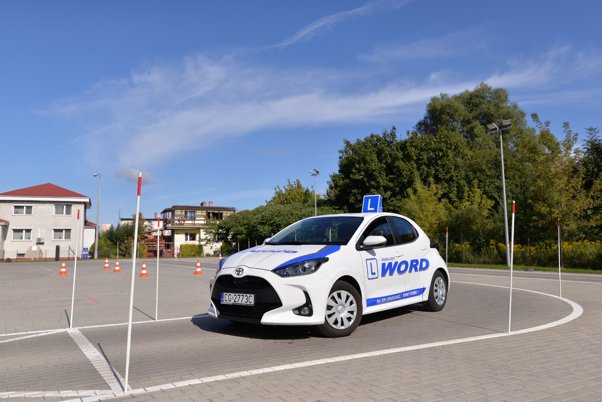 Biały samochód z niebieskim oznaczeniem 'L' i nazwą 'WORD' na dachu i boku, stojący na placu manewrowym z pachołkami i słupkami, w tle budynki i drzewa, błękitne niebo.