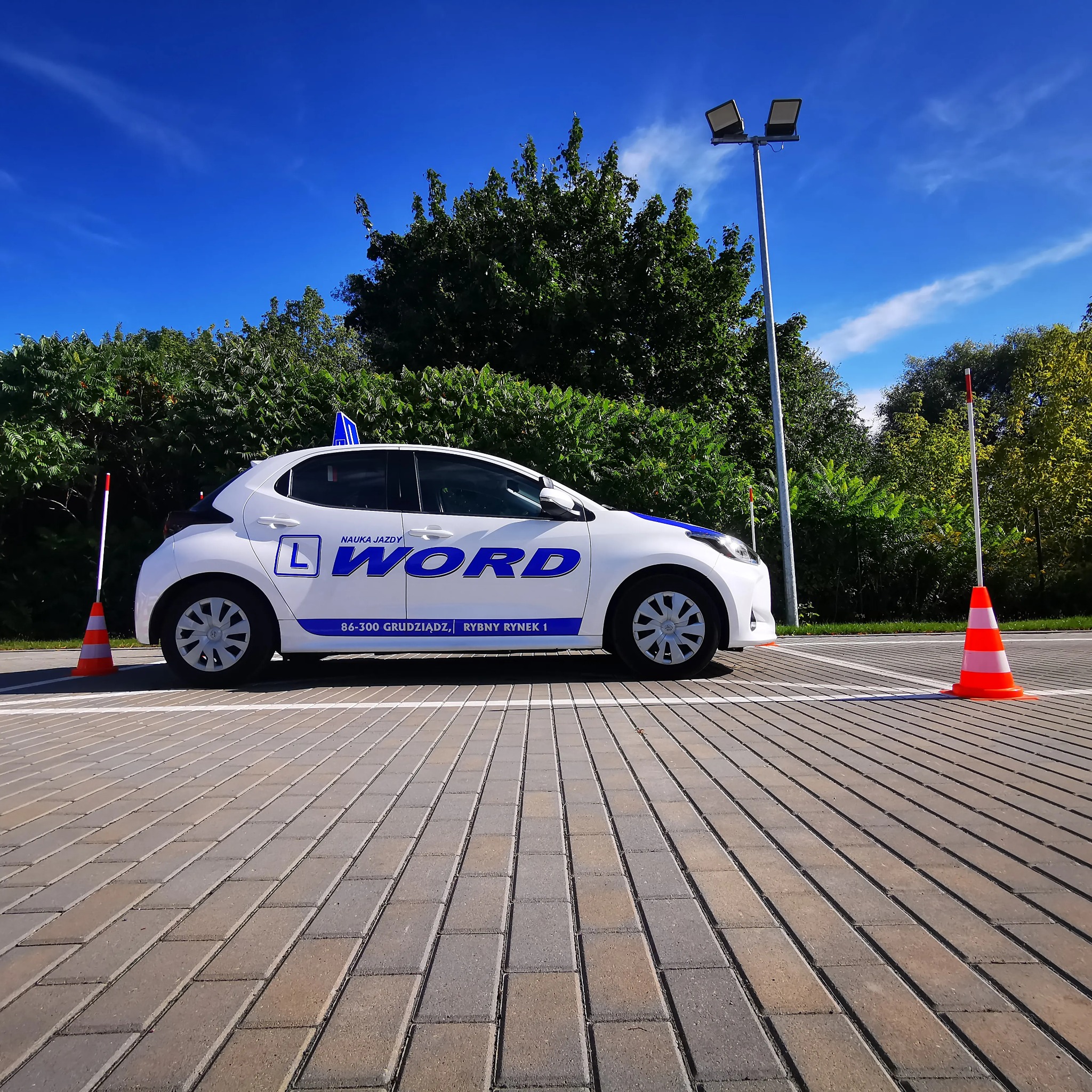 Biały samochód oznaczony literą 'L' i napisem 'WORD' na placu manewrowym z dwoma pachołkami, na tle zieleni i błękitnego nieba.