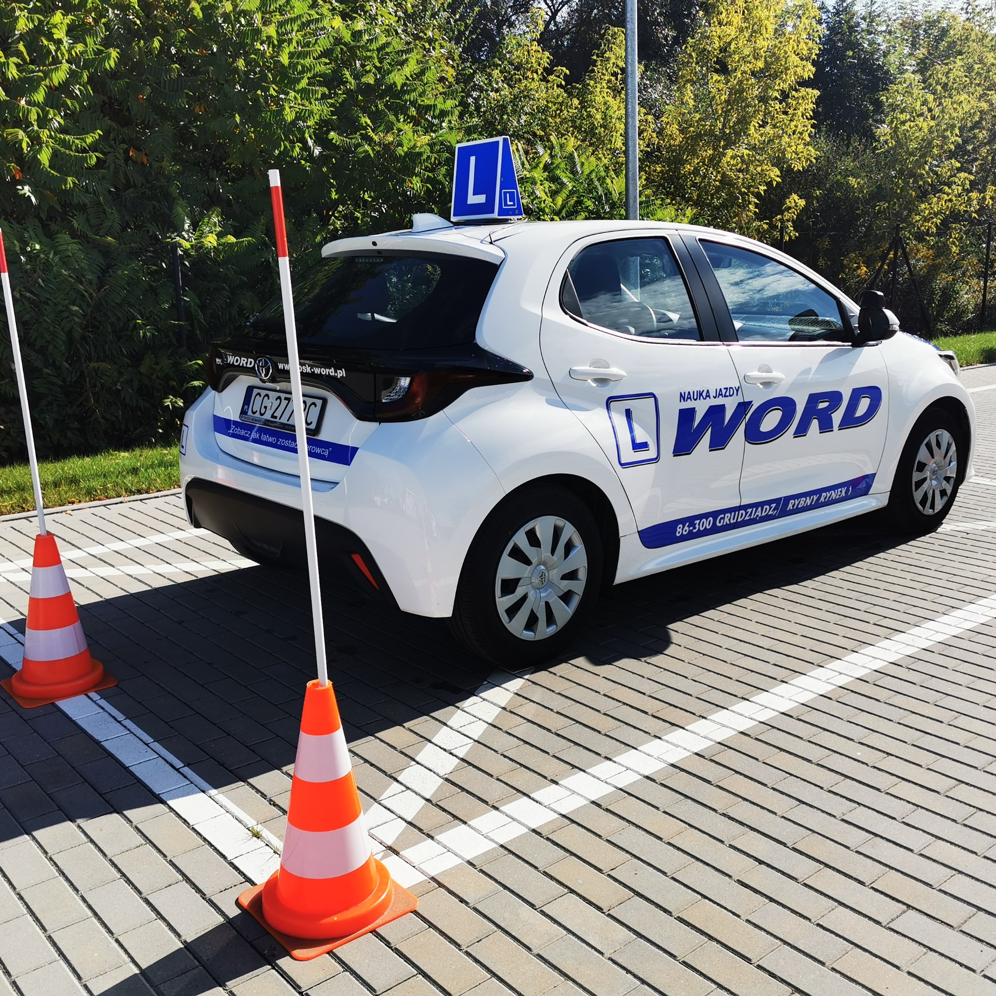 Biały samochód szkoły jazdy WORD z niebieskim oznaczeniem 'L' na dachu, zaparkowany na placu manewrowym z dwoma pomarańczowymi pachołkami i białymi słupkami.