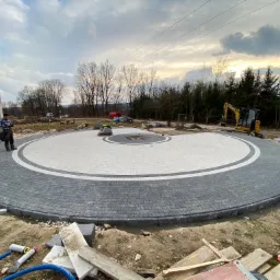 Okrągły podjazd z kostki brukowej w trakcie realizacji, z centralnym wzorem z jasnych kostek, otoczony szarym pasem i ciemniejszą obwódką. Na obrzeżach podjazdu widoczny sprzęt budowlany, w tle dom...