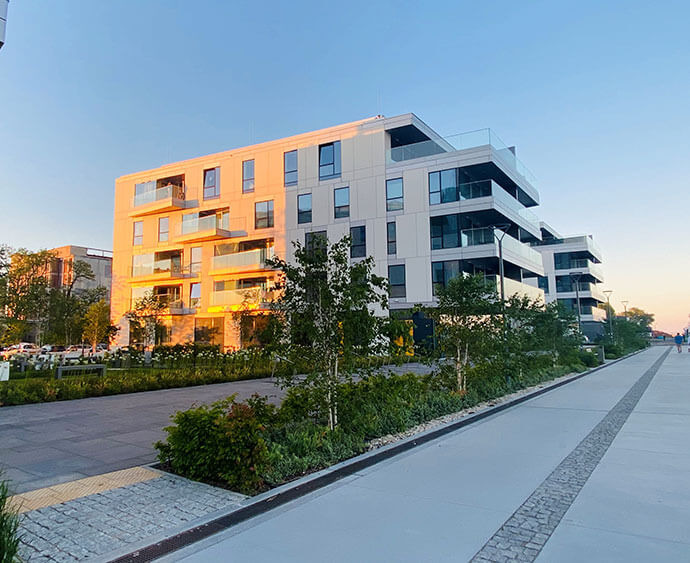 Nowoczesny budynek apartamentowy z balkonami i szklanymi balustradami, oświetlony zachodzącym słońcem, otoczony zielenią i chodnikiem.