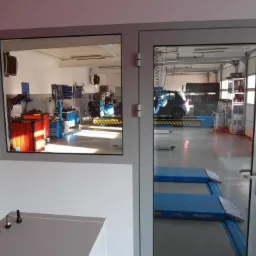 Widok przez szybę na wnętrze warsztatu samochodowego z podnośnikami, regałem z oponami i narzędziami; w tle zaparkowany samochód.