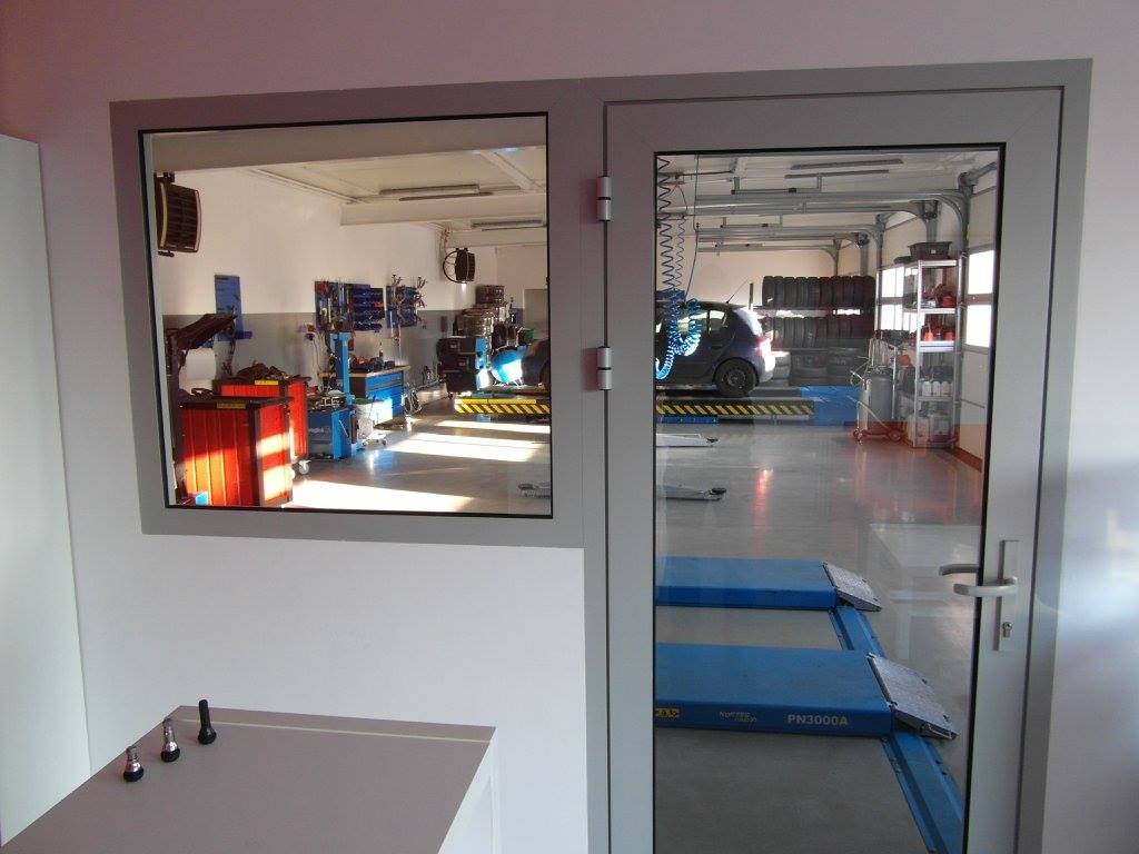 Widok przez szybę na wnętrze warsztatu samochodowego z podnośnikami, regałem z oponami i narzędziami; w tle zaparkowany samochód.