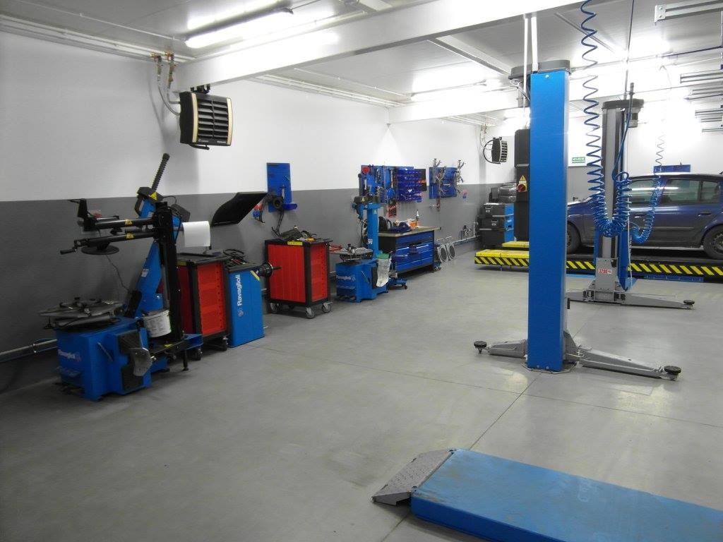 Wnętrze warsztatu samochodowego z podnośnikami, montażownicą do opon, szafkami narzędziowymi i samochodem na podnośniku.