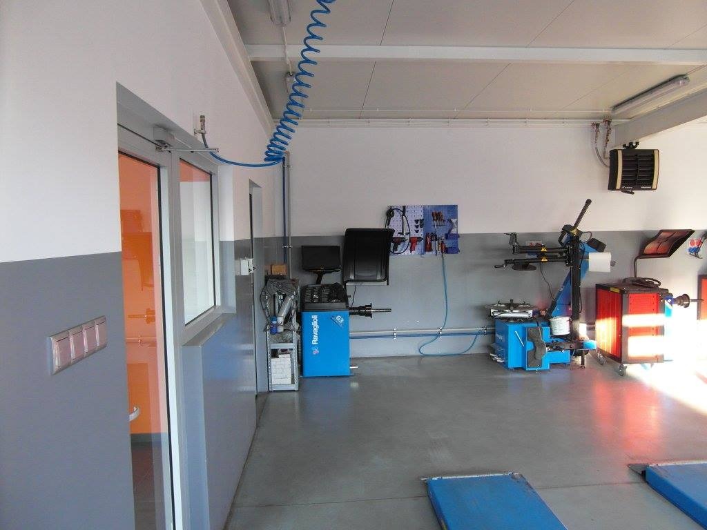 Wnętrze warsztatu samochodowego z wyposażeniem: wyważarka do kół Ravaglioli, montażownica do opon, narzędzia na ścianie i kanały najazdowe, oświetlone światłem dziennym.