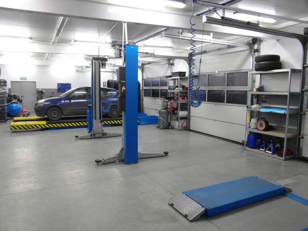 Wnętrze warsztatu samochodowego z podnośnikami, niebieskim samochodem na jednym z nich, regałami z oponami i narzędziami w tle, oraz betonową podłogą.