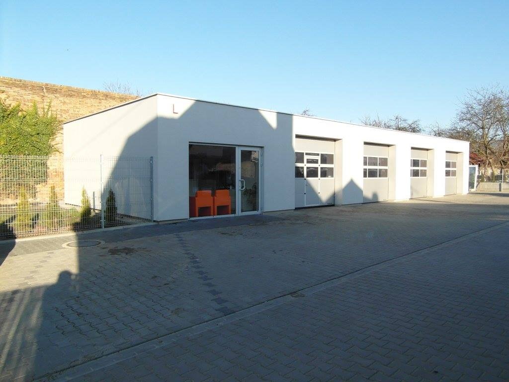 Nowoczesny budynek warsztatu samochodowego z czterema bramami garażowymi i przeszklonym wejściem, widoczny z zewnątrz, z pomarańczowymi krzesłami w poczekalni.