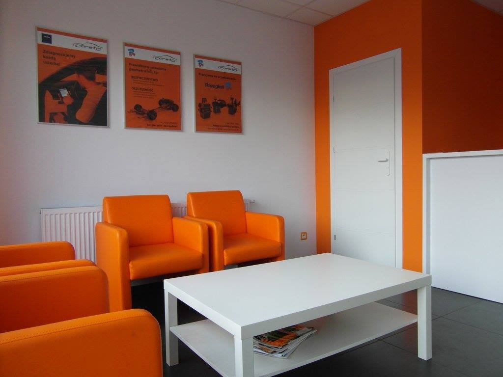Poczekalnia warsztatu samochodowego z pomarańczowymi fotelami, białym stolikiem i plakatami reklamowymi na ścianie.