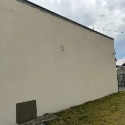 Świeżo otynkowana, beżowa elewacja budynku z widocznymi śladami po przewodach. Przy ścianie leży drabina, a w tle widoczny ogród i brama wjazdowa.