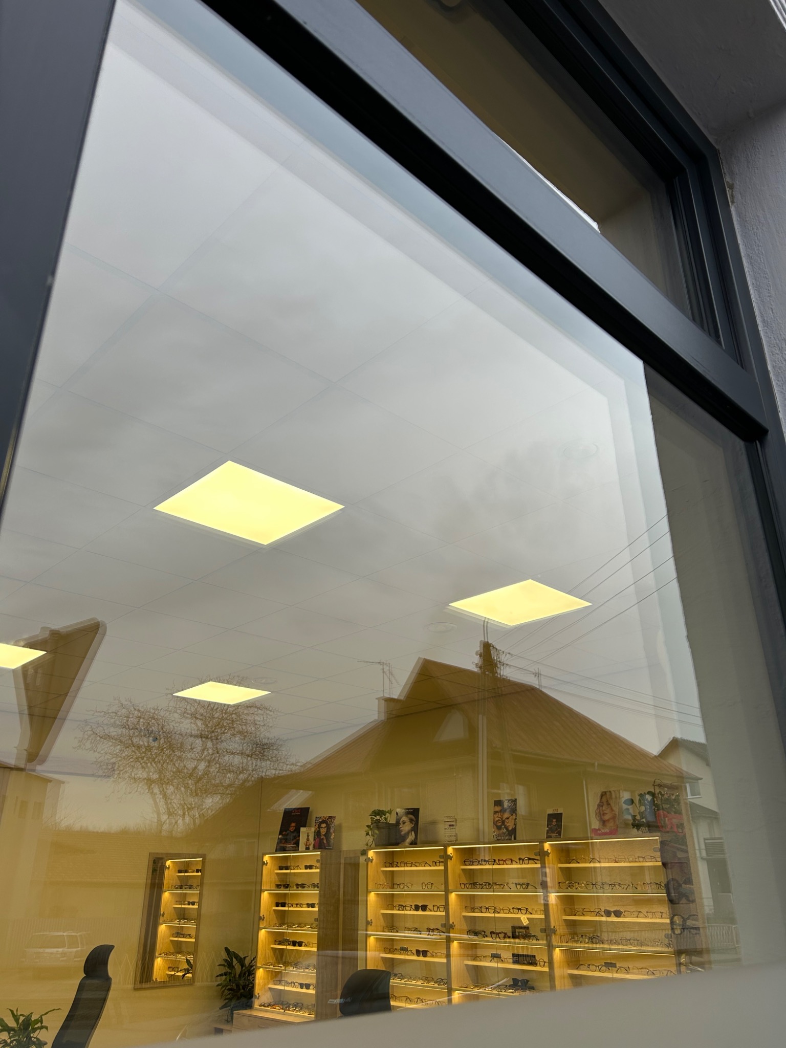 Refleksy w szybie okiennej sklepu optycznego, widoczne regały z okularami i odbicie budynku naprzeciwko.