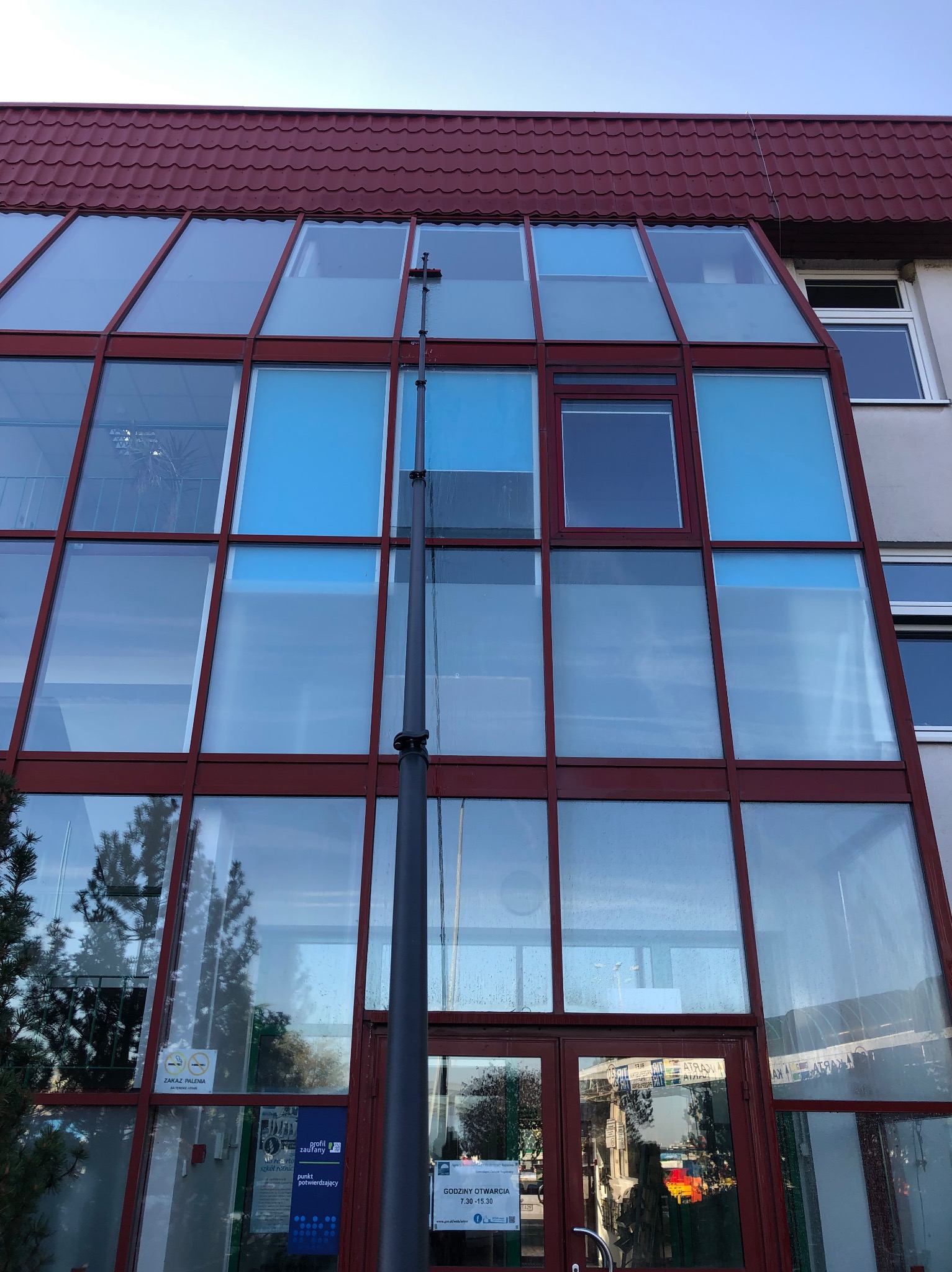 Czyszczenie szklanej elewacji budynku za pomocą teleskopowej lancy z dyszą, widoczne ślady wody na szybach, czerwona rama okienna, dach z czerwonej dachówki.
