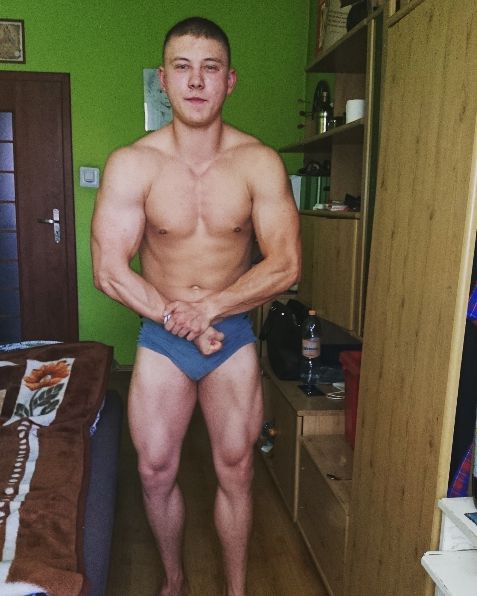 Młody mężczyzna prezentuje muskulaturę w domowych warunkach, stojąc na tle zielonej ściany i mebli z butelką wody.