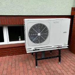 Biała jednostka zewnętrzna z wentylatorem, marki Netsu, zamontowana na czarnym stelażu z regulowanymi nóżkami, ustawiona na czerwonej kostce brukowej przed budynkiem z oknem i ceglaną podmurówką.