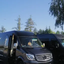 Dwa czarne busy Mercedes-Benz Sprinter, jeden z otwartymi drzwiami, zaparkowane na zewnątrz w słoneczny dzień.