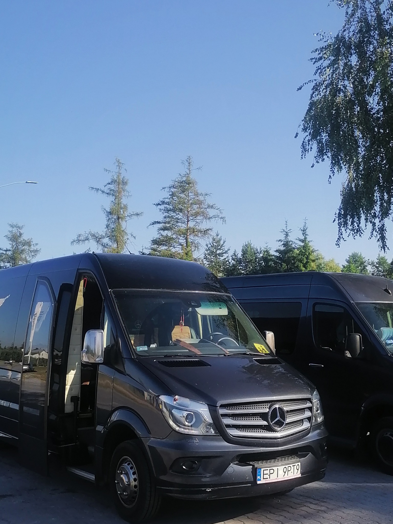 Dwa czarne busy Mercedes-Benz Sprinter, jeden z otwartymi drzwiami, zaparkowane na zewnątrz w słoneczny dzień.